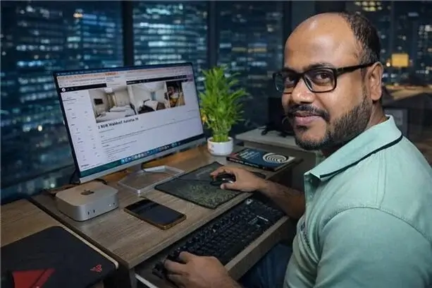 A man wearing glasses and a light green polo shirt smiles at the camera while working at a computer desk. He has one hand on a mouse and the other near a keyboard tray. On the wooden desk is a large monitor displaying a website with interior room photos, a Mac mini, a smartphone, and a small potted plant. A large window behind him reveals an illuminated city skyline at night.